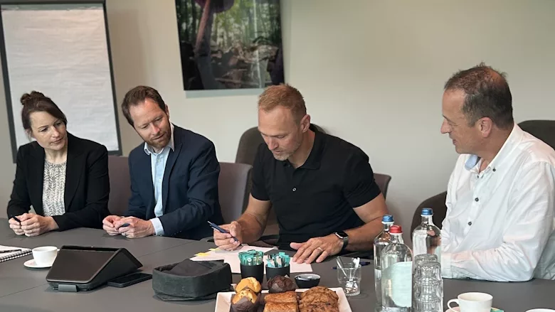 Picture of three men and a woman sitting at a table with one man signing a document